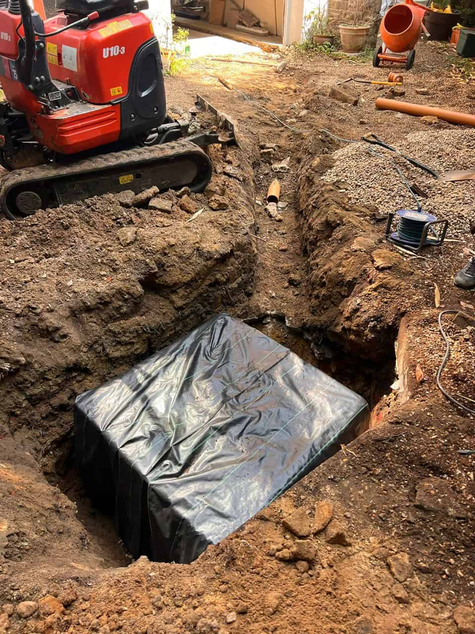Excavation for drain installation with protective plastic covering in progress.